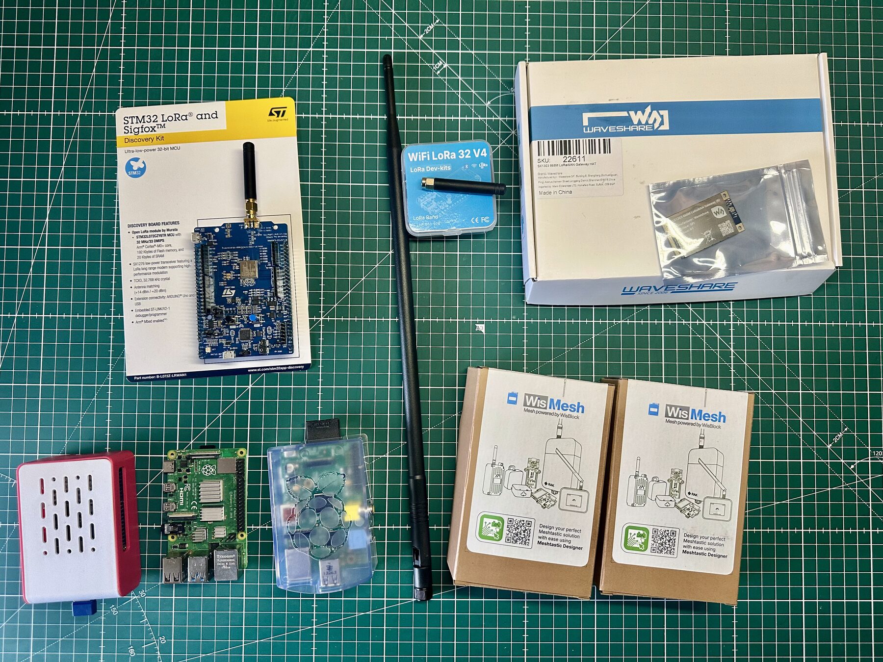 The bench laid out: STM32 Discovery kit in retail packaging, the diymore LoRa V4 board with a stub antenna, a long 868 MHz whip antenna, the Waveshare gateway HAT in its box, three Raspberry Pis (a Pi 3 in a clear plastic case, a bare Pi 4, and a Pi 5 in a red ventilated case), and two unopened WisMesh Meshtastic node boxes — all on a green cutting mat.